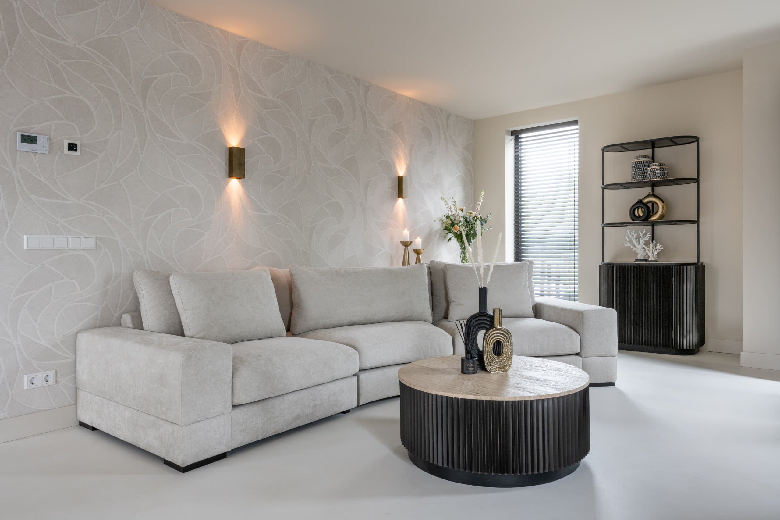 Lichte woonkamer met een crème hoekbank, een zwarte ronde salontafel met houten blad, een zwart wandrek en een zwarte vaas op tafel